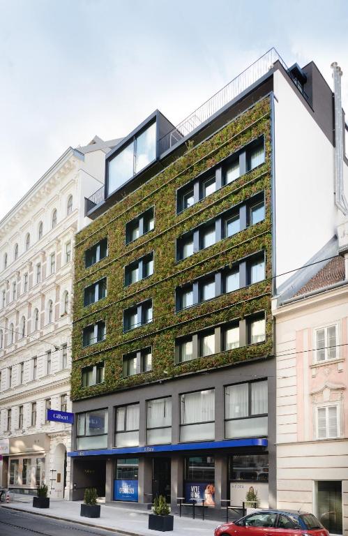 Hotel Gilbert exterior with a striking living vertical garden facade of green and flowering plants climbing between modern windows, a glass rooftop extension, and a blue entrance awning, set between ornate cream and blush Historicist buildings on a quiet cobbled street in Vienna, Austria.