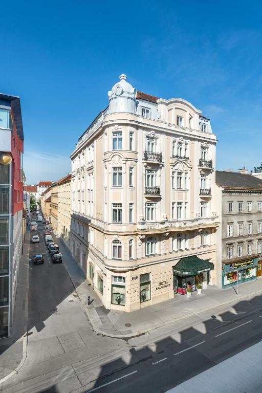 Elegant sunlit cream Historicist corner building of Hotel Johann Strauss, with a domed corner turret, wrought-iron balconies, ornate carved window surrounds, a mansard roof, and a green entrance awning with potted plants, set on a quiet sunny street under a bright blue sky in Vienna, Austria.