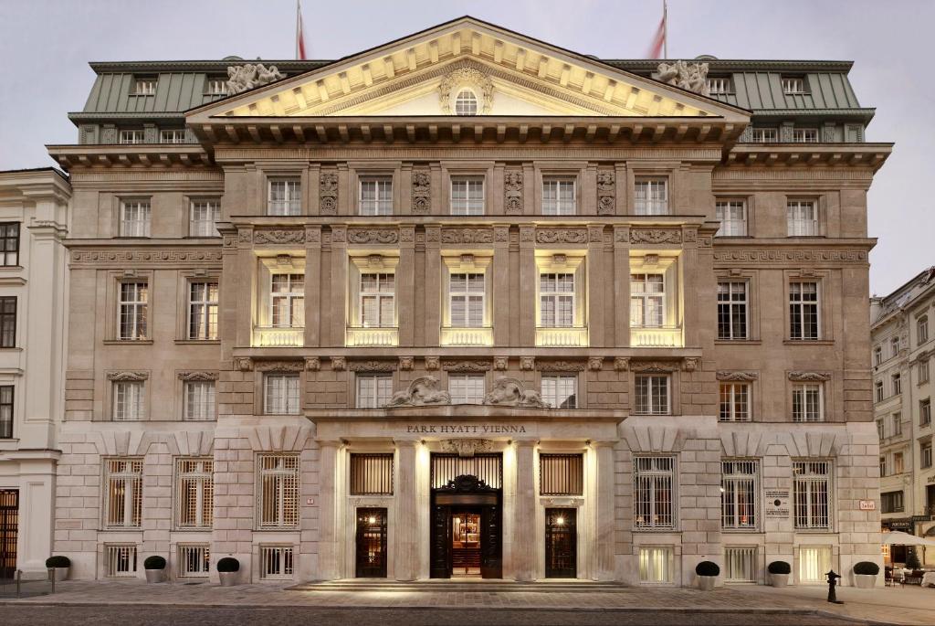 Grand Neoclassical stone facade of Park Hyatt Vienna, the former Länderbank headquarters, with a triangular pediment, carved rooftop sculptures, a columned central entrance, and warm window lights glowing at dusk on the historic cobbled Am Hof square in the Innere Stadt of Vienna, Austria.