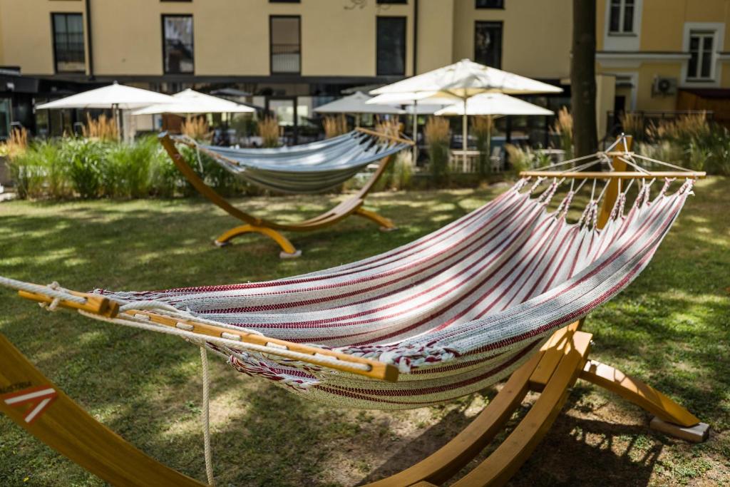 Sunny hotel garden at Austria Trend Parkhotel Schönbrunn with red-and-white and blue-striped woven hammocks on curved wooden stands across a shady green lawn, tall ornamental grasses, and white umbrellas shading the outdoor terrace of the yellow Historicist building in Vienna, Austria.