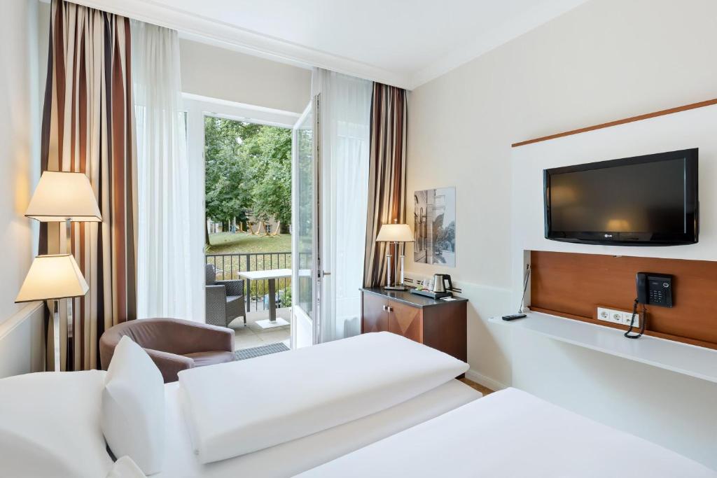 Austria Trend Parkhotel Schönbrunn guest room with twin beds in white linens, brown striped curtains, a leather tub chair, a wooden desk, a wall-mounted TV, and open French doors onto a private patio with gray wicker chairs overlooking a leafy green garden lawn and trees in Vienna, Austria.