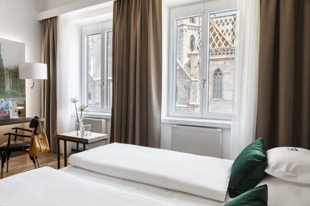 Boutique Hotel am Stephansplatz guest room with twin beds, white linens, and emerald velvet pillows, a desk with a bentwood chair and mustard handbag, and tall windows with taupe curtains framing St. Stephen’s Cathedral’s Gothic stonework and tiled roof in central Vienna, Austria.