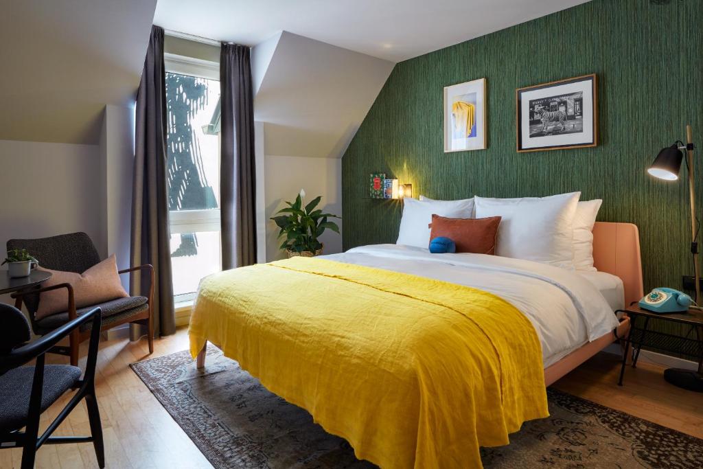 Cozy attic guest room at Max Brown Hotel 7th District with a sloped ceiling, a bed with white linens and a bright yellow linen throw, a green grasscloth accent wall with two framed prints, a vintage teal rotary phone, black floor lamp, mid-century armchair, and a rug in Vienna, Austria.