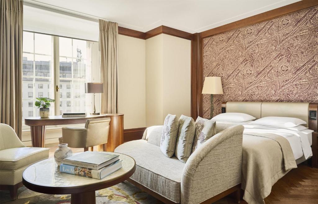 Park Hyatt Vienna guest room with a plush cream daybed sofa and embroidered pillows, a round marble coffee table holding travel books and a vase, a wood writing desk by tall windows facing historic facades, and a king bed beneath a paisley wall panel in the Innere Stadt of Vienna, Austria.