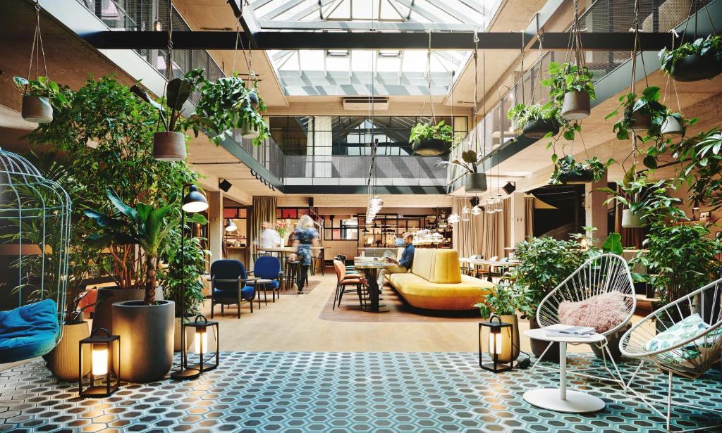 Light-filled Hotel Gilbert lobby atrium beneath a glass skylight, with hanging potted plants, leafy trees in planters, a hanging birdcage swing chair, white wire lounge chairs, glowing black lanterns, a mustard velvet banquette, and teal and blue hexagon floor tiles in Vienna, Austria.