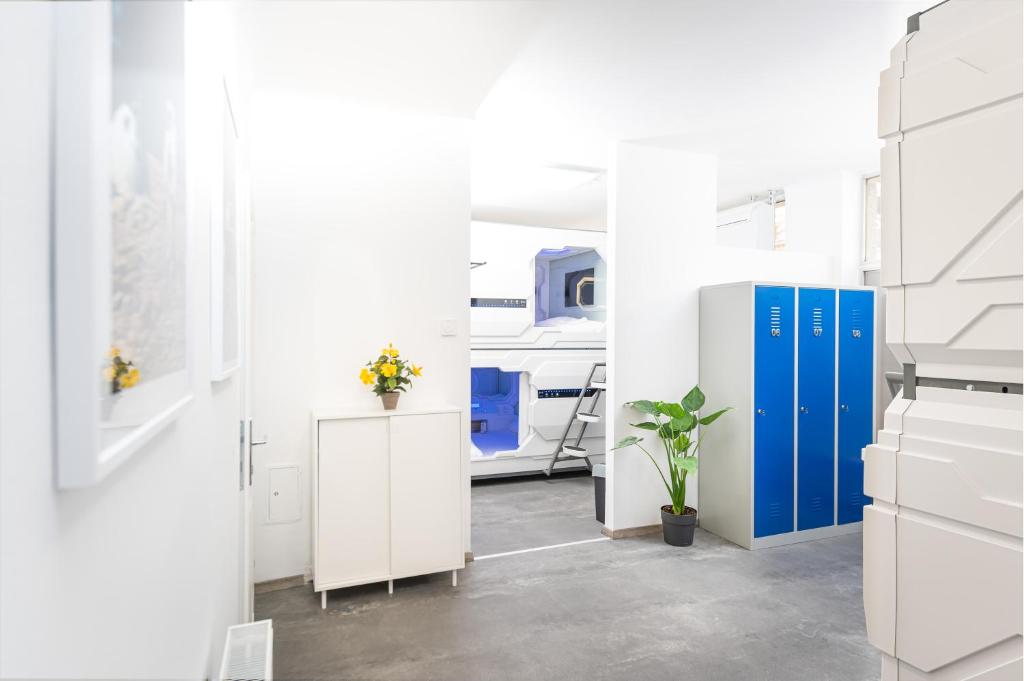 Bright white entryway at Space Home Apartment - City Center with a white cabinet topped by yellow flowers, a potted plant beside three numbered blue metal lockers, and an open doorway onto rows of futuristic white sleep capsules with blue-lit windows and a metal ladder in Vienna, Austria.
