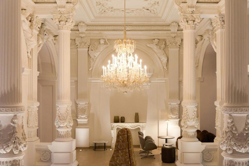 Cloud One Wien-Staatsoper hotel lobby lounge with towering fluted Corinthian columns, ornate white plasterwork, carved angel reliefs, and a sparkling crystal chandelier above a marble fireplace mantel, furnished with sleek modern swivel armchairs and a tall patterned high-back chair in Vienna, Austria.