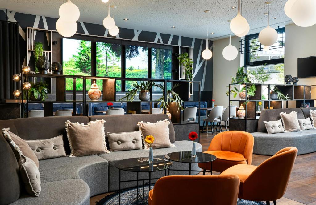 Stylish lobby lounge at Leonardo Hotel Vienna Hauptbahnhof with curved gray sofas piled with fringed pillows, orange velvet armchairs around black glass coffee tables holding single gerbera daisies, white globe pendant lights, and open shelving with plants and vases in Vienna, Austria.