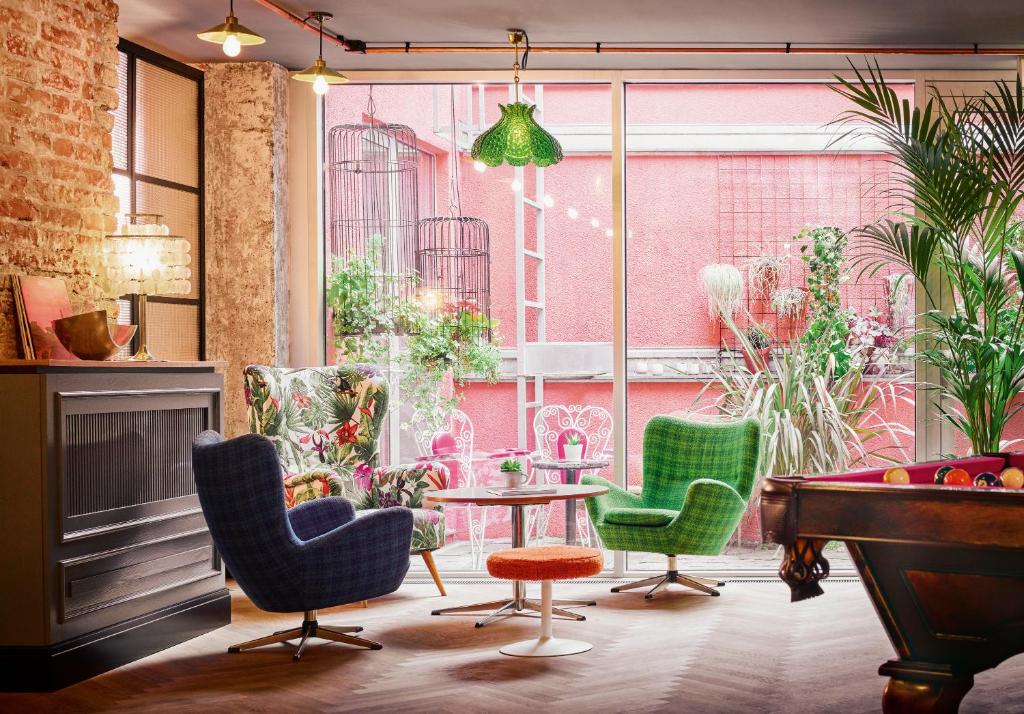 Eclectic lounge at Max Brown Hotel 7th District with retro armchairs in navy plaid, green check, and tropical floral prints around a small round table, a vintage pool table, exposed brick, a green glass pendant lamp, and windows onto a pink courtyard with birdcages in Vienna, Austria.