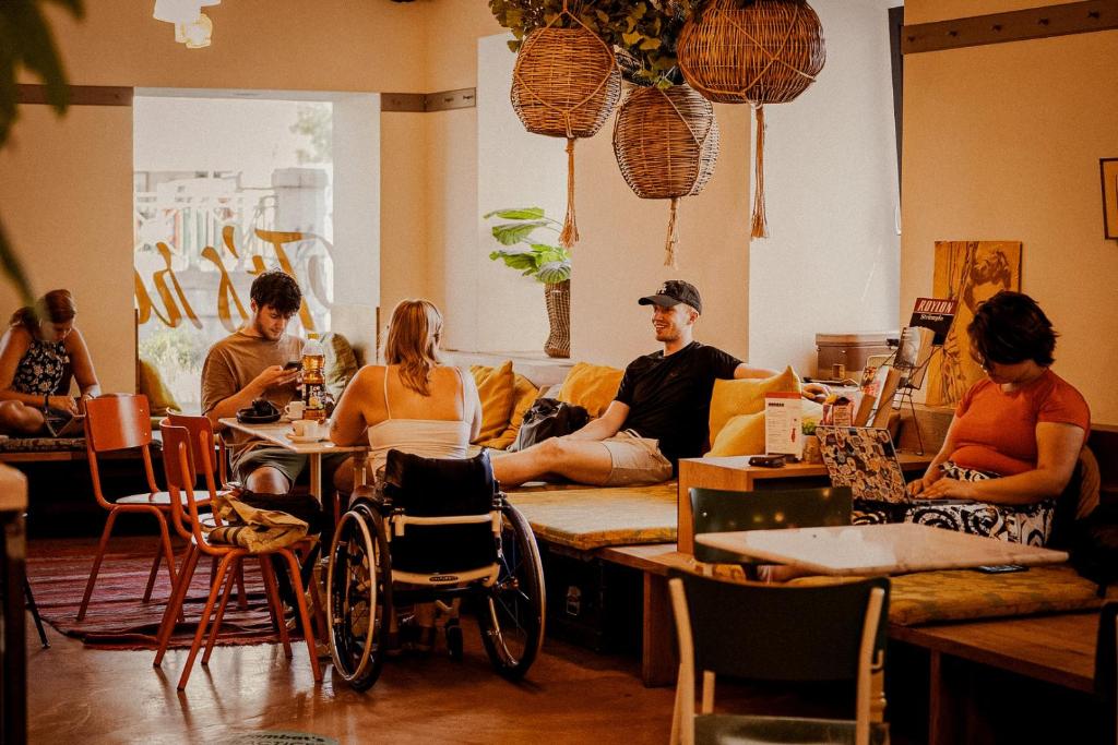 Relaxed lounge at Wombat’s City Hostel Vienna Naschmarkt with travelers lounging on mustard-cushioned benches, chatting and using laptops and phones, including a guest in a wheelchair, under woven rattan pendant lights beside potted plants and orange metal chairs and a red rug in Vienna, Austria.