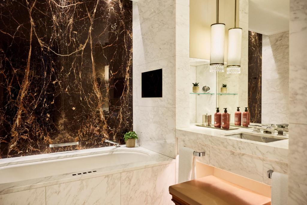Park Hyatt Vienna marble bathroom with a deep white soaking tub set against a dramatic dark brown and gold-veined marble wall, a white Carrara marble vanity with branded toiletries, twin crystal pendant lights, a wide mirror, and small potted plants in the Innere Stadt of Vienna, Austria.
