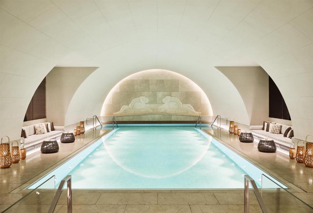 The Arany Spa indoor pool at Park Hyatt Vienna, set beneath the vaulted cream stone arches of the former bank’s lower level, with a softly glowing arched alcove and carved relief at the far end, flanked by cushioned lounge nooks, woven lanterns, and rattan side tables in Vienna, Austria.