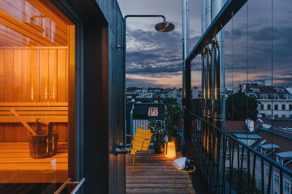 Rooftop terrace at Hotel Gilbert at dusk with a glowing wood-paneled sauna, an outdoor rain shower, a yellow metal chair, potted plants, a lantern, and folded towels, overlooking red-tiled rooftops and dormer windows beneath a purple and orange twilight sky in Vienna, Austria.