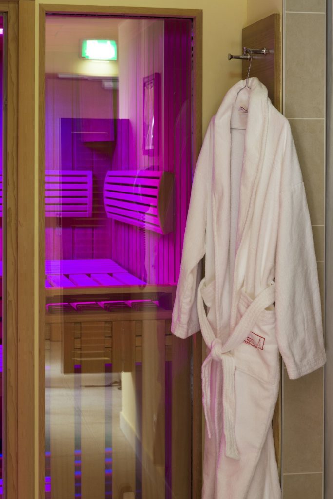 Wellness area at Boutique Hotel am Stephansplatz, with a white plush bathrobe bearing the hotel logo hanging on a hook beside the glass door of a wood-paneled infrared sauna glowing in violet and magenta light, with slatted benches and backrests visible, in central Vienna, Austria.