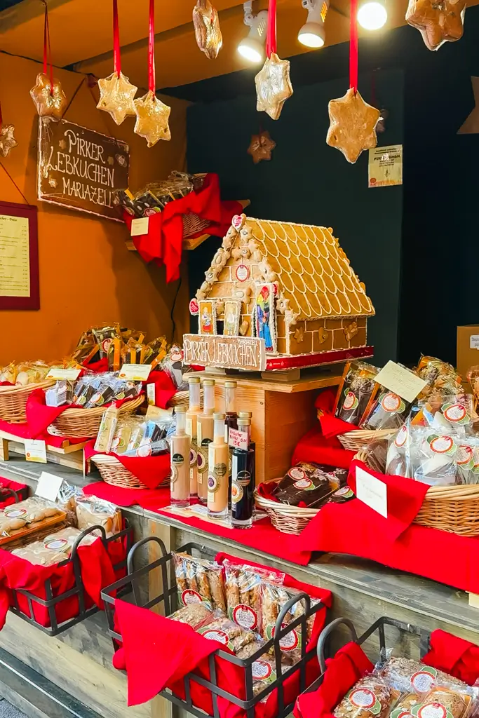 Festive Christmas market stall filled with gingerbread cookies, a decorated gingerbread house, holiday treats, and bottles displayed among bright red decorations.