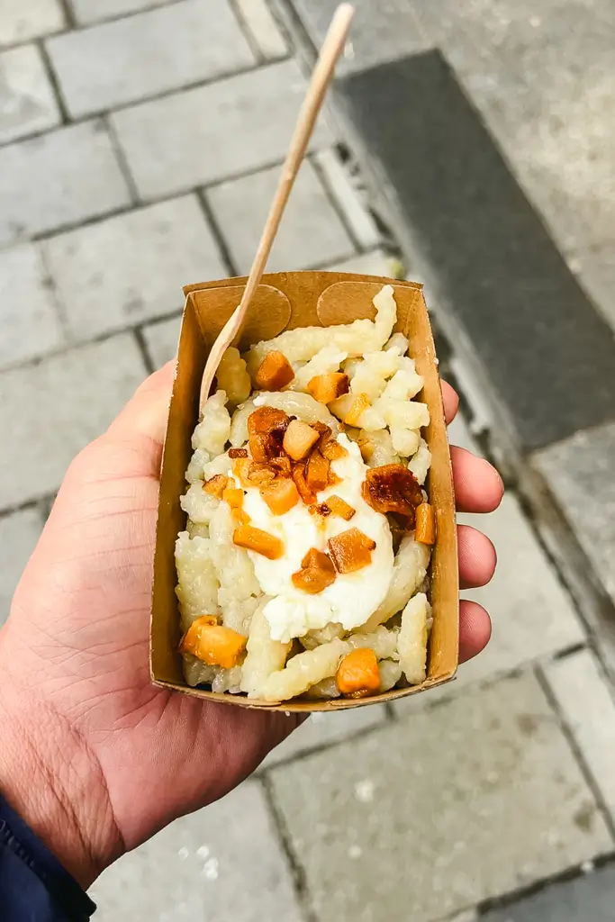Traditional halušky served in a small paper tray with creamy cheese and crispy bacon pieces at a European Christmas market.