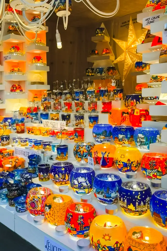 Colorful hand-painted glass candle holders featuring winter villages and festive designs displayed at a European Christmas market.