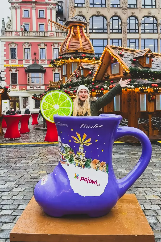 Jordyn of The Mobile Homie posing behind a giant purple mug at the Wrocław Christmas Market in Poland, surrounded by festive wooden stalls and historic architecture.