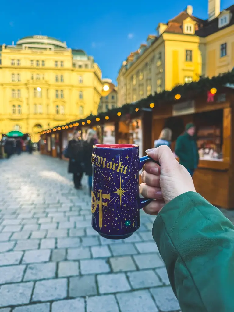 Hand holding a festive Am Hof Christmas market mug in Vienna, Austria, with wooden holiday stalls and historic buildings glowing in the background.